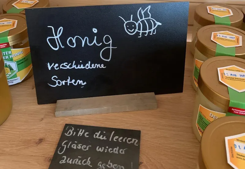Kreidetafel „Honig, verschiedene Sorten“ zwischen Honiggläsern, davor der Hinweis, leere Gläser zurückzubringen